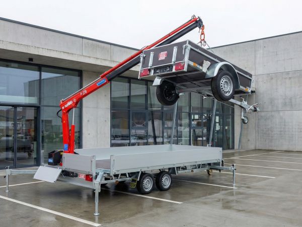 Ein silberner Tandemanhänger mit ausgefahrenen Stützfüßen und einem roten Ladekran, der gerade einen kleineren Pkw-Anhänger in die Luft hebt