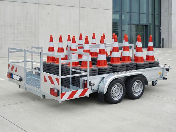 Ein silberner Tandemanhänger mit rot-weißer Warnmarkierung, beladen mit mehreren Stapeln rot-weißer Leitkegel zur Absicherung von Baustellen