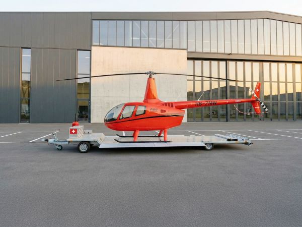 Roter Hubschrauber auf einem silbernen Transportanhänger vor einem modernen Industriegebäude im warmen Abendlicht.
