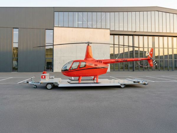 Roter Hubschrauber auf einem silbernen Transportanhänger vor einem modernen Industriegebäude im warmen Abendlicht.