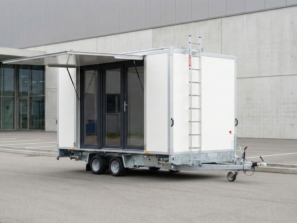 Moderner, weißer Büroanhänger auf einem Tandemfahrgestell mit großen Glastüren, geöffnetem Vordach und seitlicher Leiter vor einer Industriehalle
