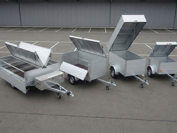 Vier silberne Aluminium-PKW-Anhänger verschiedener Größen mit geöffneten Deckeln, die nebeneinander auf einem gepflasterten Platz stehen.