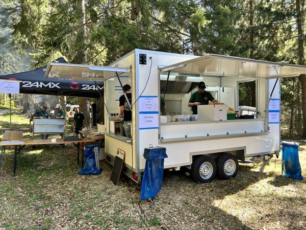 Weißer Verkaufsanhänger mit aufgeklappten Klappen beim Outdoor-Event im Wald