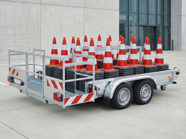 Ein silberner Tandemanhänger mit rot-weißer Warnmarkierung, beladen mit mehreren Stapeln rot-weißer Leitkegel zur Absicherung von Baustellen