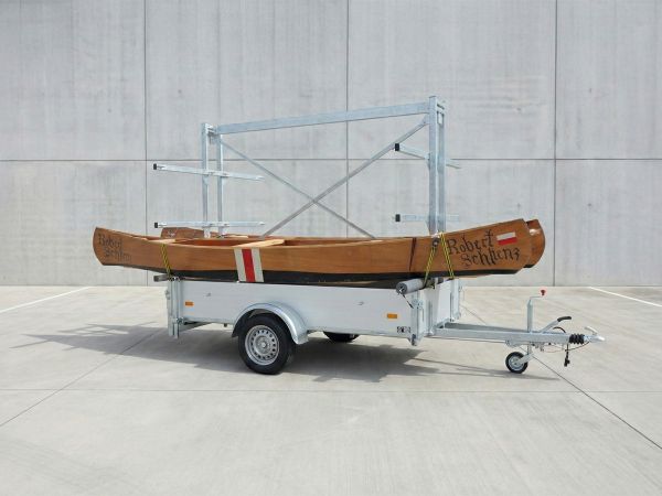 Bootsanhängers mit Kastenaufbau und hohem verzinkten Metallgestell, auf dem ein braunes Holzkanu geladen ist.