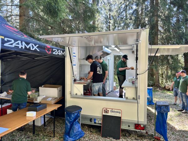Verkaufsanhänger im Outdoor-Einsatz bei einem Event: Team bereitet Speisen zu, daneben ein Pavillon im Wald