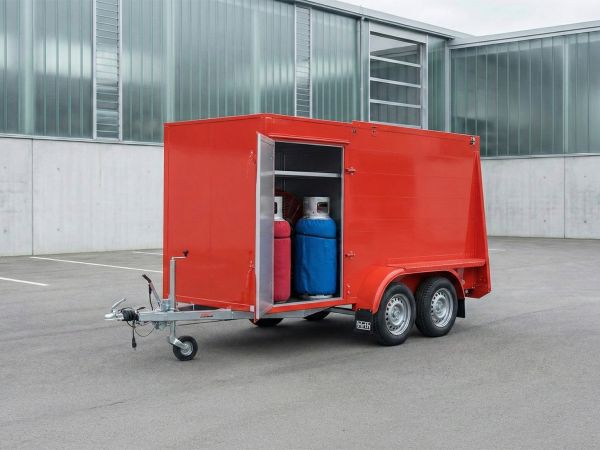 Ein leuchtend roter Ballonanhänger mit geöffneter Seitentür und sichtbarem Equipment steht auf einem sauberen Asphaltplatz vor einer modernen Industriehalle mit Glasfassade.