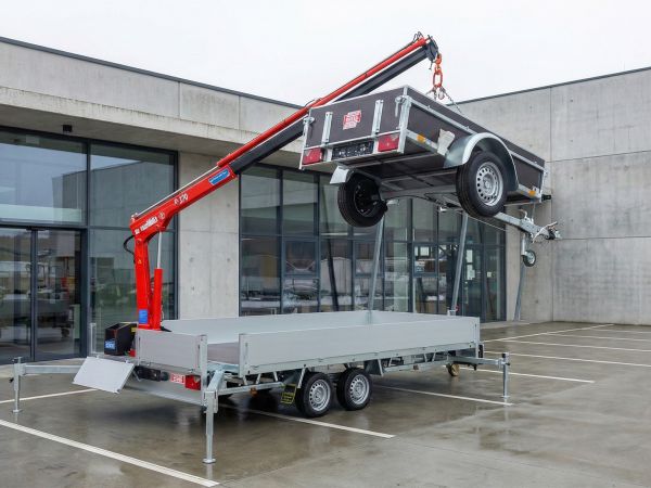 Ein silberner Tandemanhänger mit ausgefahrenen Stützfüßen und einem roten Ladekran, der gerade einen kleineren Pkw-Anhänger in die Luft hebt